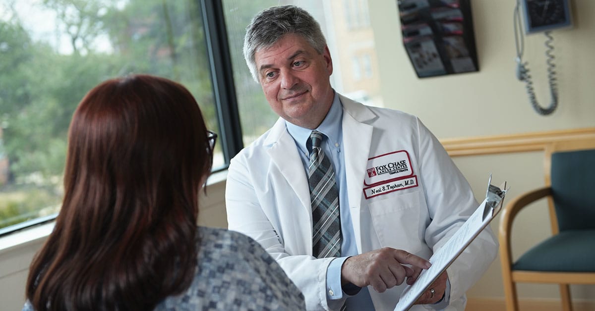 A photograph of a Fox Chase doctor sitting across from a patient, smiling as he points to something on a clipboard.